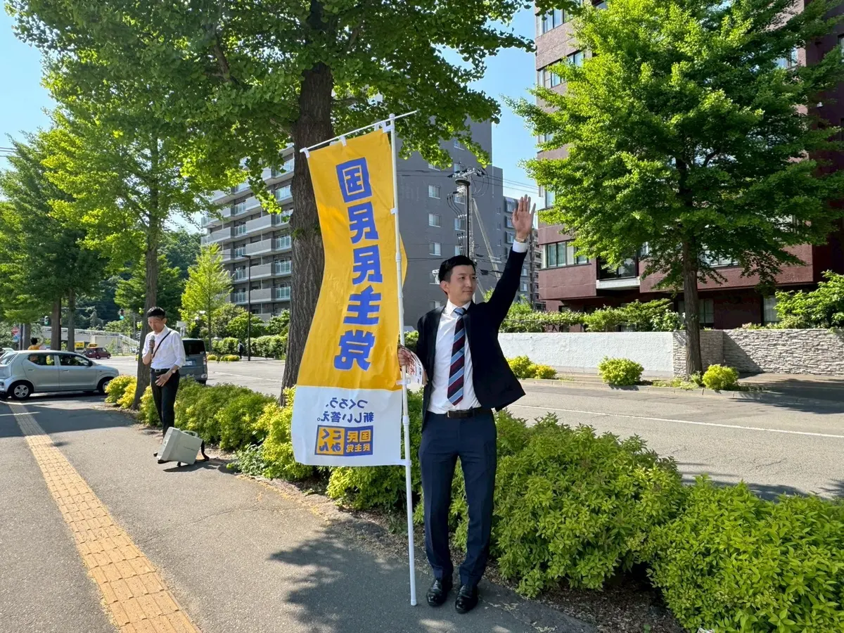 街路樹の並ぶ通りで旗を掲げて手を振る阿部恭介